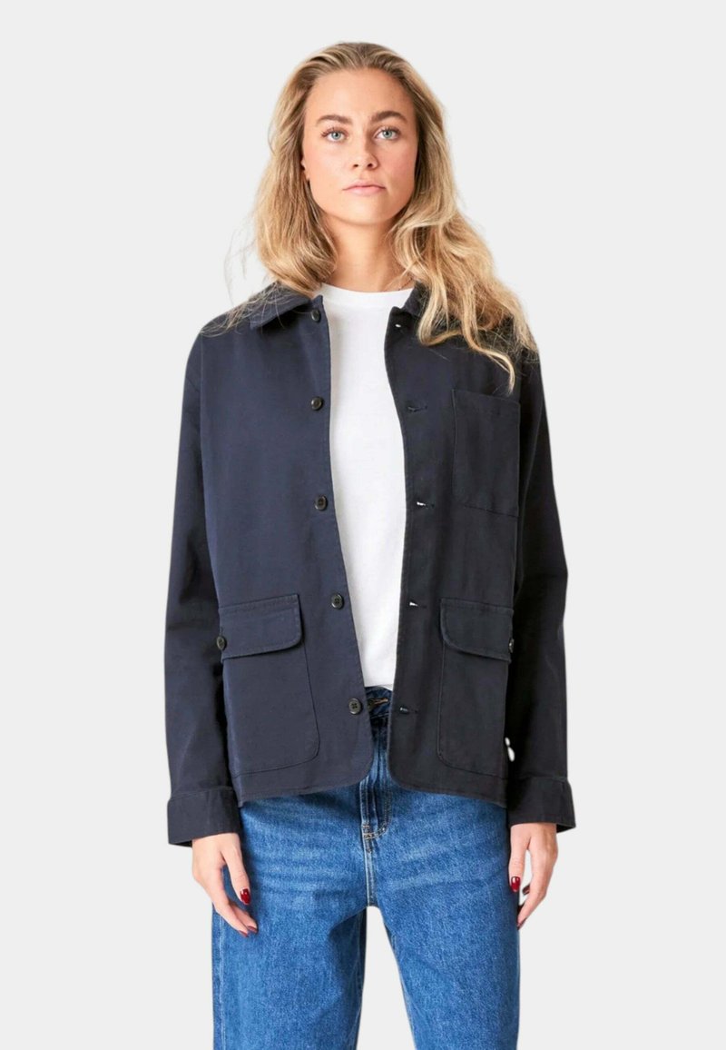 Marineblå jakke med krave, knappelukning foran og to påsyede lommer. Fremstillet af et glat, holdbart stof, parret med en hvid skjorte.