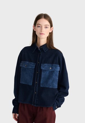 Jeune femme aux longs cheveux portant une chemise bleue foncée boutonnée avec de grandes poches poitrine et un pantalon marron, devant un fond uni.