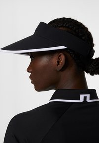 Svart skärm med en bred brätte, med en vit accent. Den justerbara remmen är av elastiskt material och visas från sidan mot en neutral bakgrund.