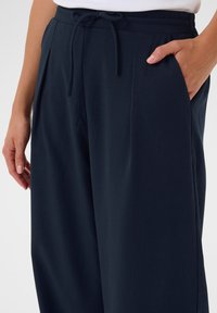 Dunkelblaue Hose mit lässiger Passform, featuring einen Kordelzugbund, Falten und seitlichen Taschen. Glatte Stoffstruktur und taillierte Gestaltung.