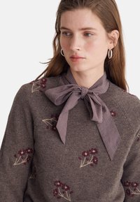 Maglione marrone lavorato a maglia con ricamo floreale in toni rossi. Colletto con papillon a quadri come dettaglio. Texture liscia e vestibilità rilassata.