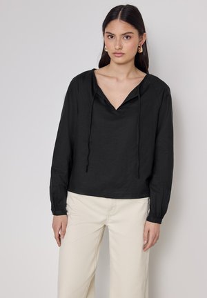 Black, long-sleeve shirt with a v-neck and adjustable drawstrings, made from lightweight fabric. Model wears cream-colored pants.
