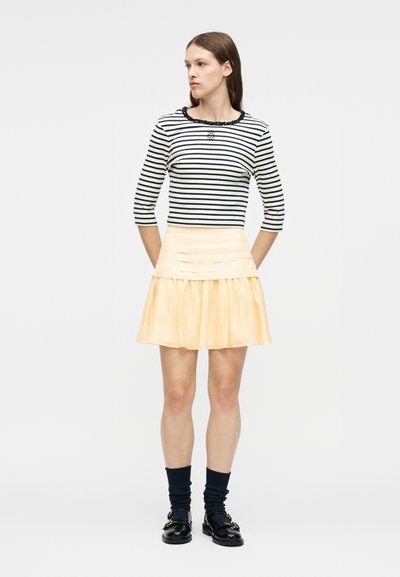 Γυναίκα όρθια με τα χέρια πίσω από την πλάτη, φοράει μαύρο και άσπρο ριγέ τοπ, ανοιχτό κίτρινη φούστα, μαύρες κάλτσες και μαύρα παπούτσια.