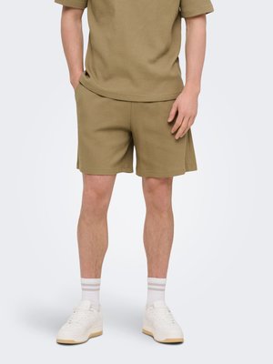 Man draagt olijfgroene, gestructureerde korte broek en bijpassend kort mouw shirt met witte sokken en witte sneakers tegen effen achtergrond.