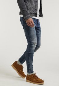 Jeans ajustados azules con detalles desgastados, combinados con zapatos de ante color beige y calcetines grises, rematados con una chaqueta de denim gris sobre una camisa blanca.