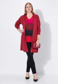 Rode lange cardigan met een open voorkant en zakken, gelaagd over een rood-zwart gradient top, gecombineerd met zwarte leggings en hoge hakken.