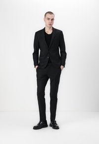 Schwarzer maßgeschneiderter Anzug mit einem Blazer mit einem Knopf, schmal geschnittenen Hosen und einem schwarzen T-Shirt darunter. Aus glattem Stoff mit klassischem Design.