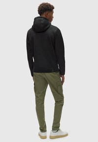 Felpa nera con cappuccio e finitura testurizzata, abbinata a pantaloni cargo verde oliva con tasche laterali e polsini aderenti. Sneakers bianche casual.