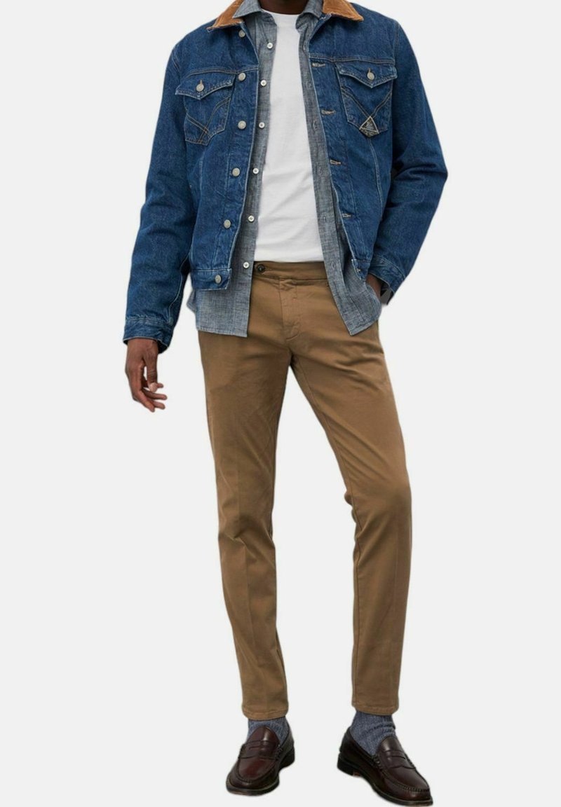 Homme portant une veste en denim bleu avec un col marron, une chemise grise, un t-shirt blanc, un pantalon beige, des chaussettes grises et des mocassins marron foncé, debout avec une main dans la poche.