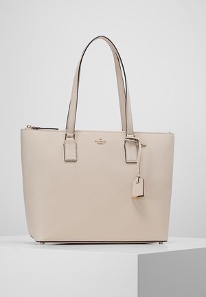 Beżowa skórzana torba typu tote z podwójnymi uchwytami, górnym suwakiem i małą zawieszką, umieszczona na białym postumencie na jednolitym tle.