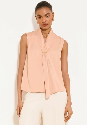 Kobieta w bezrękawowej morelowej bluzce z plisą z przodu i złotą agrafką, zestawiona z białymi spodniami o wysokim stanie, stoi na jednolitym tle.