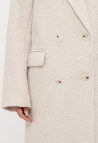 Διπλής όψεως παλτό σε απαλό μπεζ υφασμάτινο σχέδιο, με μεγάλους κρεμ κουμπιά και μια πλαϊνή τσέπη στα αριστερά.