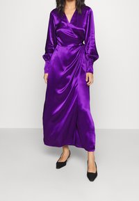 Lila Satin-Wickeldress mit langen Ärmeln, tiefem V-Ausschnitt und seitlichem Bindeband. Glatte Textur und fließende Silhouette. Kombiniert mit schwarzen Schuhen.