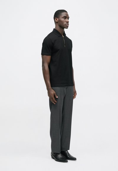 Juodas polo marškinėlis su užtrauktuku ant apykaklės, derinamas su pilkomis kelnėmis ir juodais odiniais batais. Lygus audinys, aptemptas dizainas ir subtilios detalės.