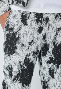 Gros plan sur un pantalon à motif floral noir et blanc avec une main dans la poche gauche et une partie d'une chemise blanche visible.