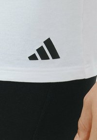 Valkoinen urheilupaita lyhyillä hihoilla ja musta adidas-logo helmassa. Kontrastiset musta ja valkoinen värit korostavat muotoilua.