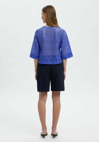 Blå, kortärmad, öppenstickad topp med rundad halsringning, kombinerad med mörka shorts. Tyget har en mönstrad, ventilerande textur.