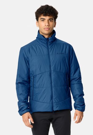 Giovane uomo con capelli corti e ricci che indossa una giacca blu con cerniera e pantaloni neri, in piedi davanti a uno sfondo semplice.