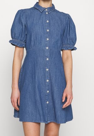 Femme portant une robe en denim bleu à manches courtes bouffantes, col avec boutonnière sur le devant.