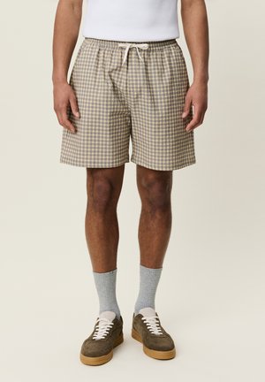 Mann trägt beige-braune karierte Shorts mit weißem Kordelzug, graue Socken und braune Sneakers und steht vor neutralem Hintergrund.