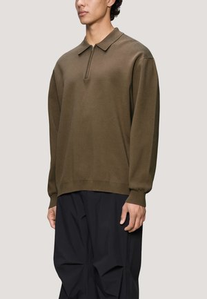 Jeune homme portant un polo manches longues vert olive avec une courte fermeture éclair et un pantalon noir ample, debout sur un fond clair.
