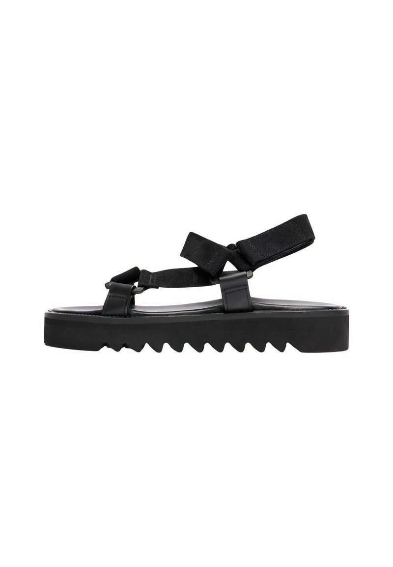 Crne sandale s debelim potplatom sa uzorkom cik-cak, podesivim trakama i glatkim gornjim materijalom. Uključuju čičak trake.