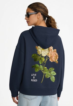 Vrouw met een zonnebril en een marineblauwe hoodie met een grote roos en de woorden "LOVE & ROSES" op de rug, van achteren gezien.