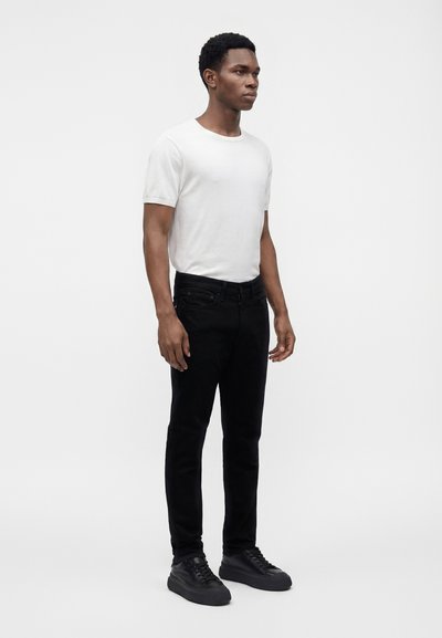 Homme debout, face à droite, portant un t-shirt blanc uni à manches courtes, un pantalon noir et des baskets noires sur fond blanc.