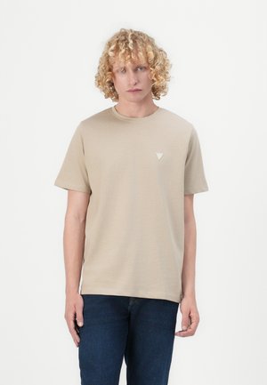 Beige bomulds t-shirt med rund hals og korte ærmer, med et lille trekantet logo på venstre bryst. Bæret sammen med mørke denim jeans.