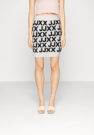 Kobieta ubrana w szarą dzianinową spódnicę z czarnym wzorem liter "JJXX", różowy krótki top i białe klapki na obcasie z odkrytymi palcami, stojąca na jednolitym jasnym tle.