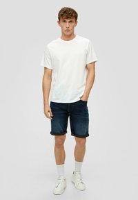 Biała koszulka z krótkim rękawem, ciemne dżinsowe szorty z podwiniętymi mankietami, białe sneakersy i szare skarpetki do kostek. Prosty, casualowy design bez wzorów.
