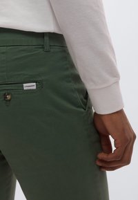 Πράσινο βαμβακερό παντελόνι με λείο ύφασμα, μία πίσω τσέπη και διακριτική επωνυμία. Το μοντέλο φοράει λευκό πουκάμισο με μακριά μανίκια.