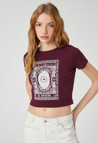 Bordeaux beskåret t-shirt med hvid grafisk design, der har indviklede mønstre og tekst. Korte ærmer og ribbet tekstur.