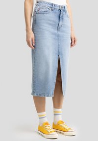 Jupe en denim bleu clair, coupe crayon, avec une fente à l'avant, un design à cinq poches et des passants de ceinture. Portée avec des baskets jaunes et des chaussettes rayées.