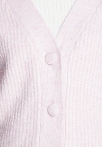 Vaaleanpunainen neuletakki, joka on valmistettu teksturoidusta kankaasta. Siinä on V-aukko, kaksi pyöreää nappia ja edessä olevat resorikuviot.