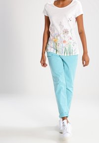 Kobieta w białej koszulce z kwiatowymi i motylkowymi nadrukami, jasnoniebieskich spodniach i białych sneakersach idzie do przodu.