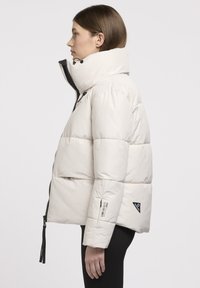 Veste doudoune beige clair avec un col montant, fermeture éclair et étiquettes d'accent. Présente des sections matelassées et un extérieur brillant et lisse.