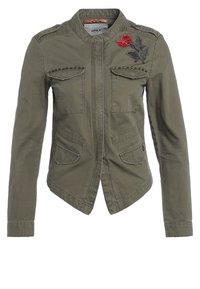 Olive zielona kurtka wojskowa w stylu z dwoma kieszeniami na piersi, z kolcami i haftowanym wzorem kwiatowym na ramieniu. Teksturowany materiał.