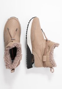 Un par de botas de tobillo beige con cuero texturizado, forro de pelo sintético, suelas de cuña y cordones decorativos sobre un fondo blanco.