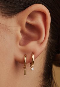 Des boucles d'oreilles en or en forme de créole présentent une finition lisse, l'une avec un charme rectangulaire et l'autre ornée d'une petite perle blanche.