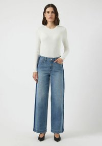 Top blanco de manga larga con canales, ligero, combinado con jeans de mezclilla azul de pierna ancha que presentan paneles laterales en contraste y una cintura alta.