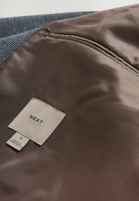 Tejido de seda marrón con finas rayas verticales, que presenta una etiqueta que dice "NEXT" y "M", junto a un panel texturizado azul.