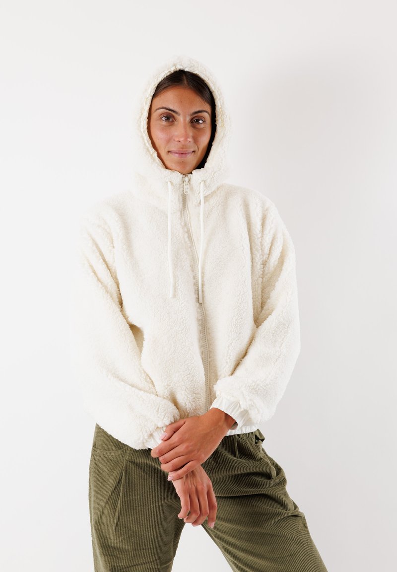Flauschige weiße Kapuzenjacke mit Reißverschluss, elastischen Bündchen und Kordelzügen, kombiniert mit olivgrünen Cordhosen.