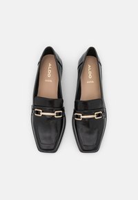 Svarta lädermockasiner med en glänsande yta, fyrkantig tå och guldtonad kedjedekoration. Fjädrande innersulor, märkta "ALDO" på bruna insidor.