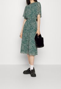 Vihreä kukkakuosiset mekko, jossa on liehuvat hihat, istuva vyötärö ja polvipituinen hame. Yhdistetty mustan tekoturkishandbagin ja paksupohjaisten kenkien kanssa.