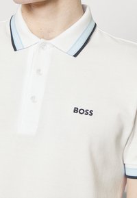 Bílá polo košile s texturovanou látkou. Obsahuje tříknoflíkovou légu, kontrastní límec s černými a světle modrými pruhy a vyšitým logem "BOSS".