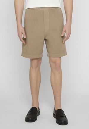 Mann iført beige knelange shorts med oppbrettede kanter, hvit skjorte og svarte lærloafers foran en ensfarget grå bakgrunn.
