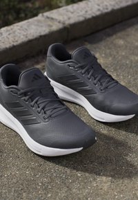 Chaussures de course Adidas gris foncé avec semelles blanches placées sur un trottoir extérieur près d’un bord en pierre.