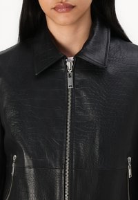 Svart skinnjakke med krokodillemønster, med høy krage, sølvfarget glidelås og glidelåslommer på siden.