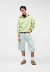 Ανοιχτό πράσινο fleece πουλόβερ με μισό φερμουάρ και λογότυπο, συνδυασμένο με ανοιχτό μπλε τζιν σορτς. Καφέ παπούτσια και λευκές κάλτσες ολοκληρώνουν το look.